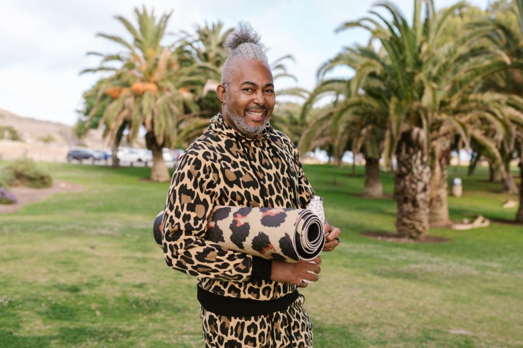 Senior man in animal print outfit holding yoga mat in sunny park, smiling joyfully.